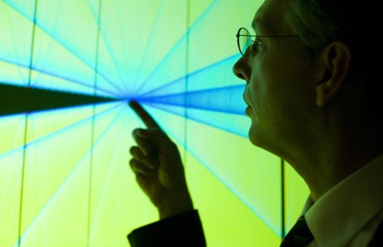 A researcher points his index finger at a screen that is displaying a colorful, radiating visualization.
