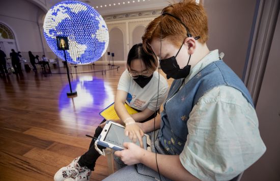 Two people sitting in chairs looking at a tablet with a large lit-up globe displayed in the background.