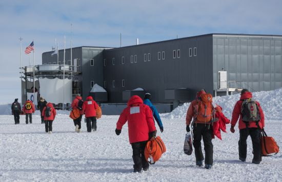 People in red jackets carrying backpacks and duffel bags towards a building in the snow.
