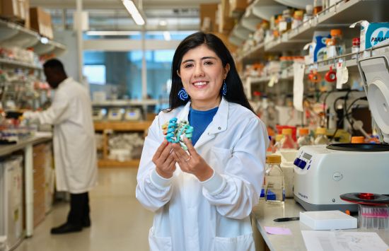 A researcher in a white lab coat stands at a lab bench holding a small blue model of a protein.