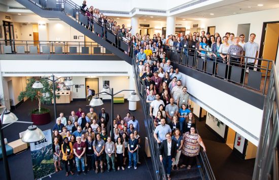 A large group of individuals, spread out across stairs, a mezzanine, and the ground floor. Next to the group on the ground floor is a pop-up banner for the NSF Environmental Data Science Innovaiton and Inclusion Lab (ESIIL).