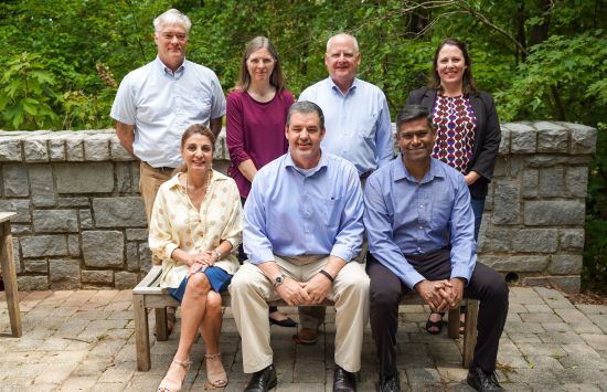 Seven researchers, four men and three women, are lined up in two rows. The first row is seated on a bench with the others standing behind them.