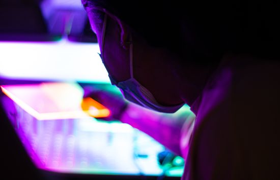 A semi-profile view of a masked researcher holding a glowing rectangular object in a dimly lit room.