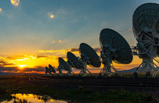 A series of massive telescope dishes all pointing the same direction against an orange-blue sky at dusk.