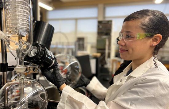 A researcher in a lab coat and safety glasses adjusting glass lab equipment.usts large