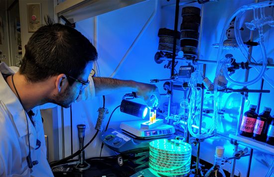 A chemist manipulates equipment in a fume hood lit with blue light.