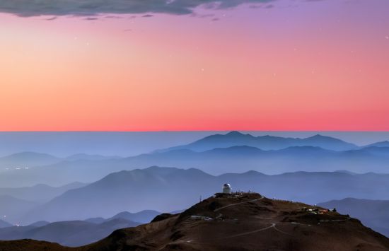 A colorful sunset and layers of blue mountaintops backdrops the Cerro Tololo Inter-American Observatory. A small profile image of the Víctor M. Blanco 4-meter Telescope is visible atop Cerro Tololo itself.