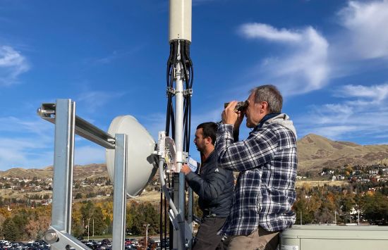 Two people, one with binoculars, stand on some equipment in front of a hillside under a bright blue sky.