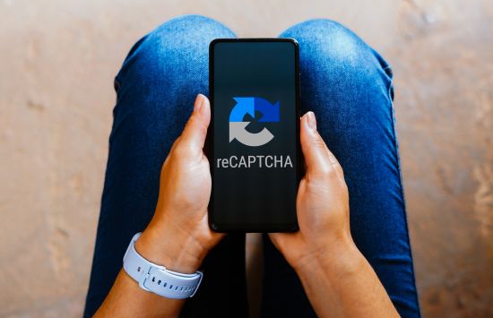 A person holds a smart phone in their lap, which displays the word "RECAPTCHA" on its screen.