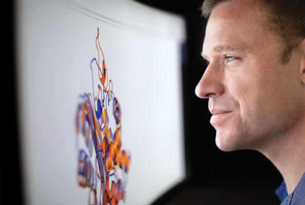 A man smiles slightly as he stares at a large computer screen that displays a multicolored model of a virus