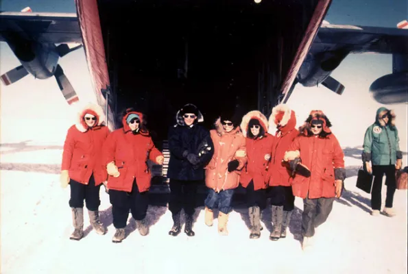 Six women in cold weather gear stand in the snow with a large aircraft behind them