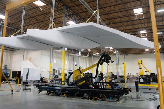 A white aircraft wing suspended above a mobile robot with a large yellow arm in an open hangar space.