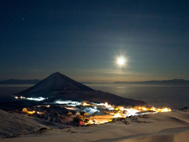 The moon shines in a dark sky over a snowy valley with small buildings illuminated by yellow light