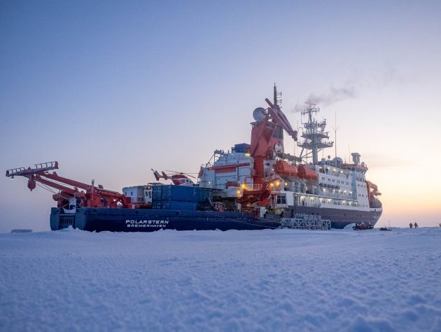 A large ship is seen in the background with a large sheet of snow and ice surrounding it