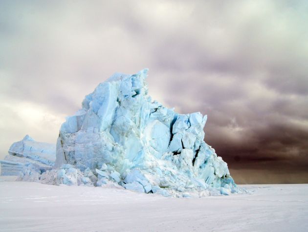 A large, whitish blue and  jagged  iceberg juts out of the flat white ground. To the right, a small figure in a red coat is seen.