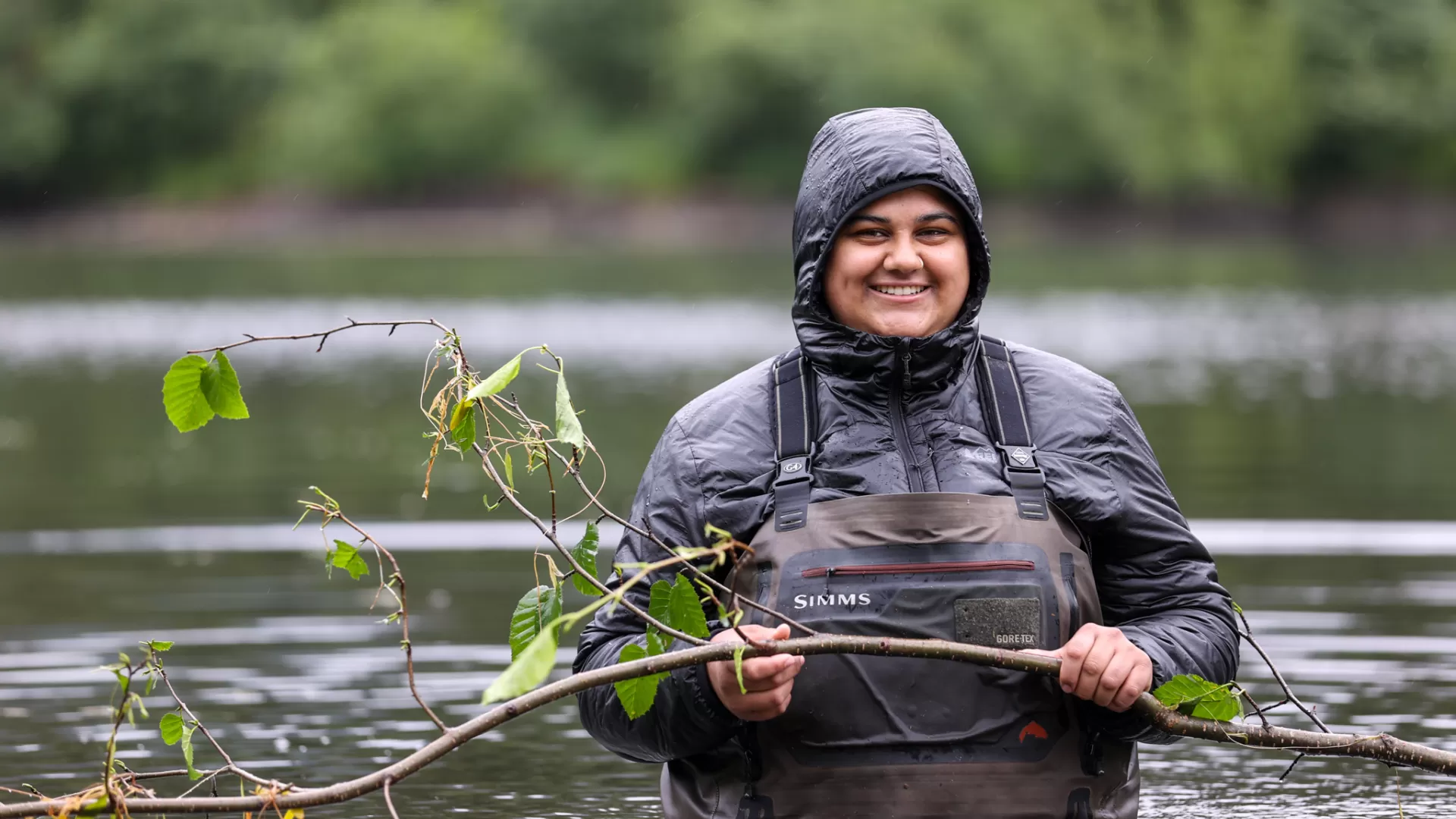 A smiling woman, wearing overalls and a hood, holding a tree branch and standing waist-deep in a river.