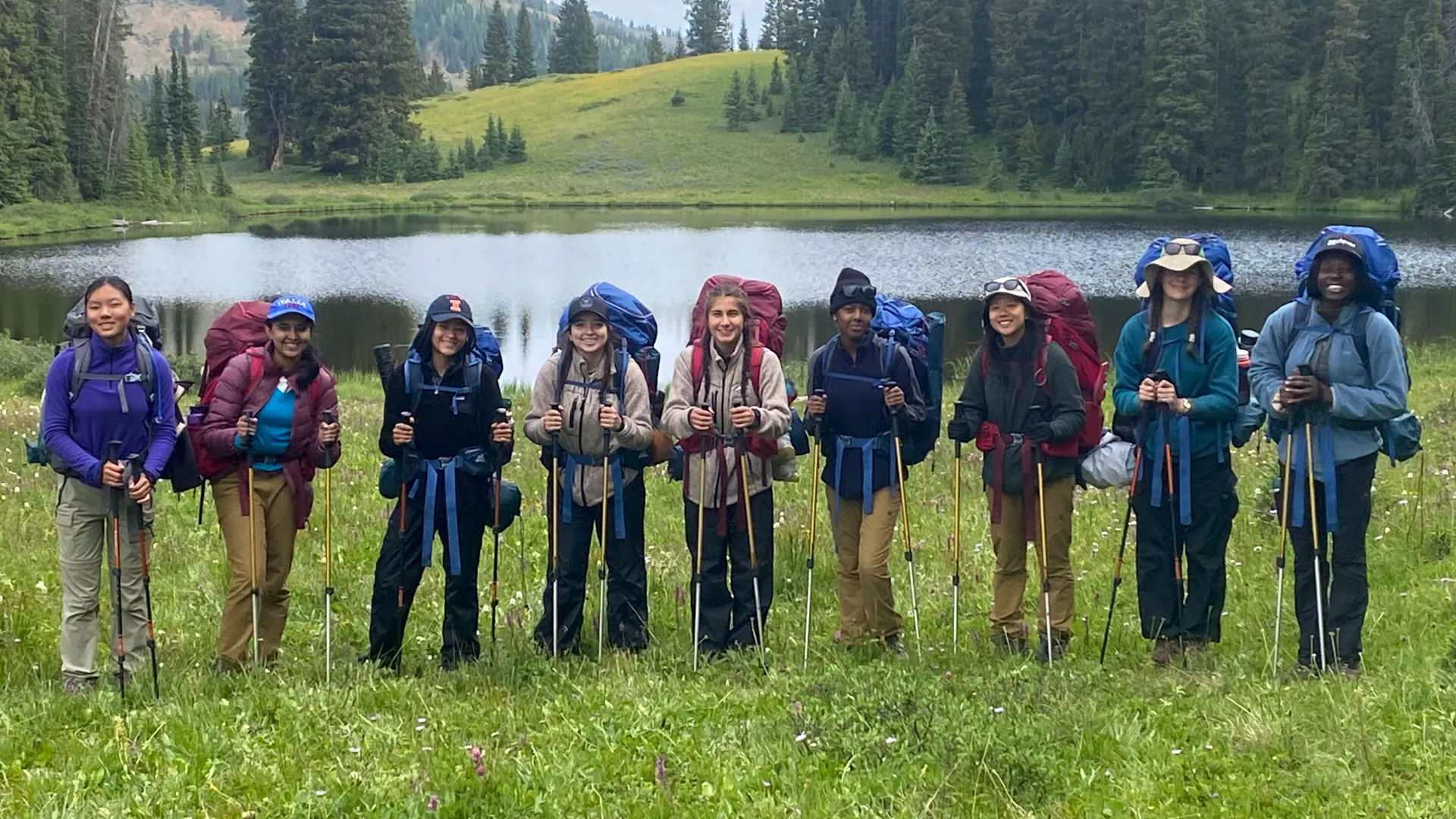 A line of smiling students with large backpacks and hiking poles posing in a meadow in front of a lake.