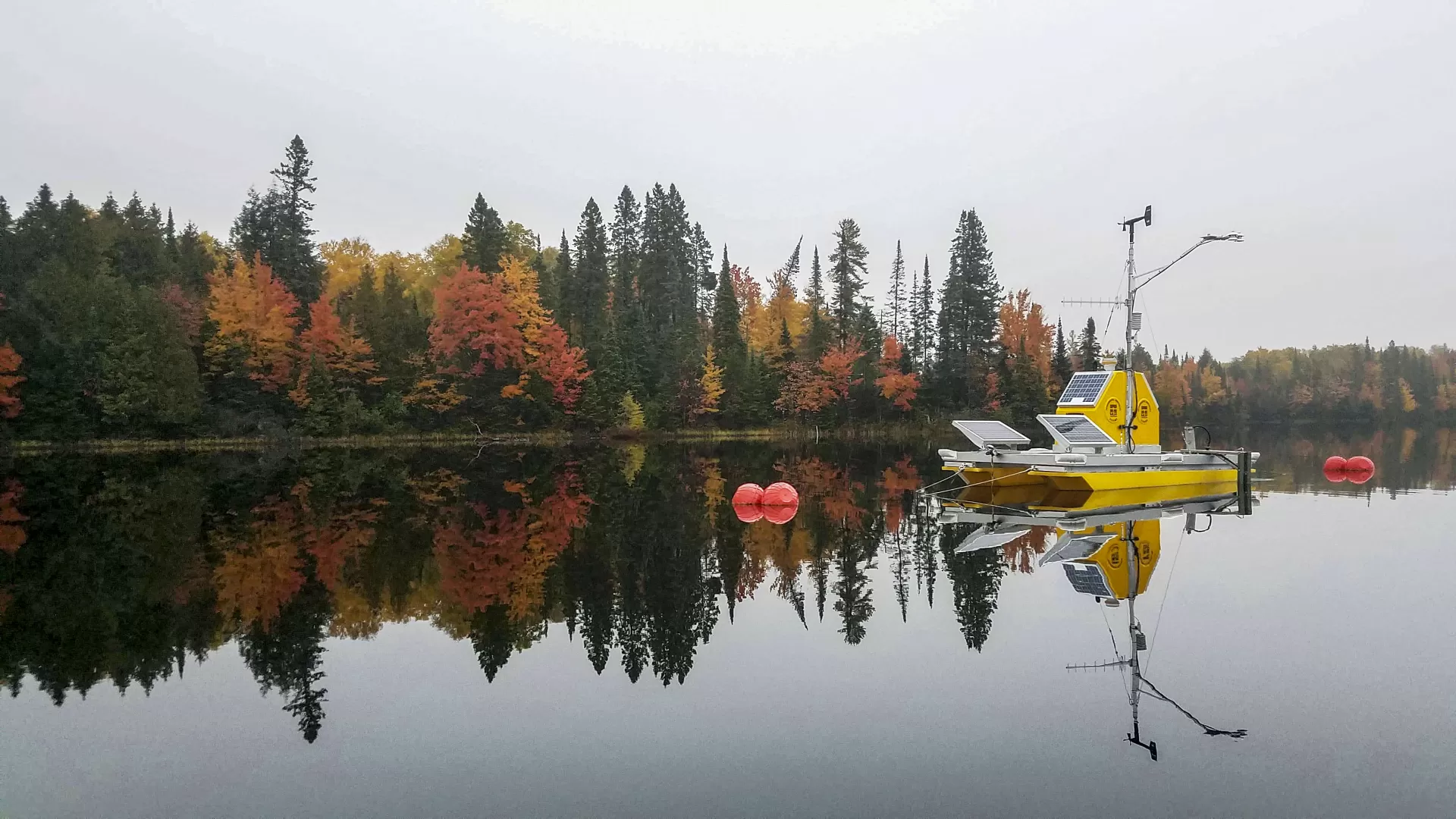 A yellow platform with solar panels and other equipment on it floating on a still lake with trees showing fall colors in the background.