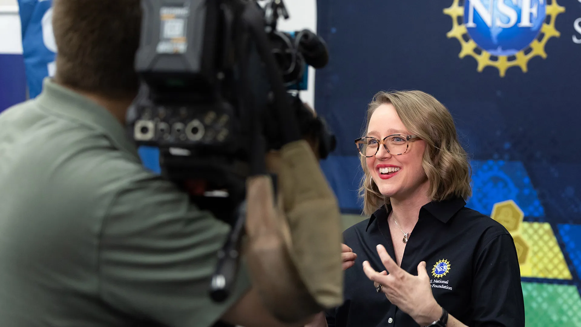 A scientist being filmed by a news camera while speaking in front of an NSF banner.