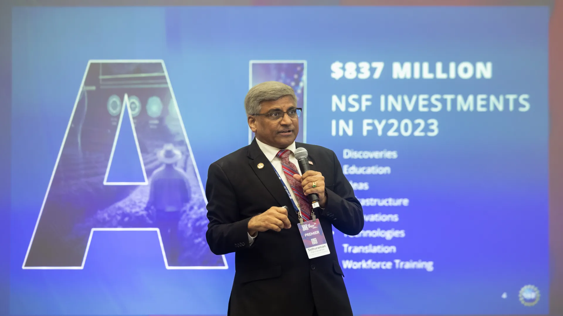 The NSF Director speaks into a microphone against a background display that states "AI: $837 million: NSF investments in FY2023"