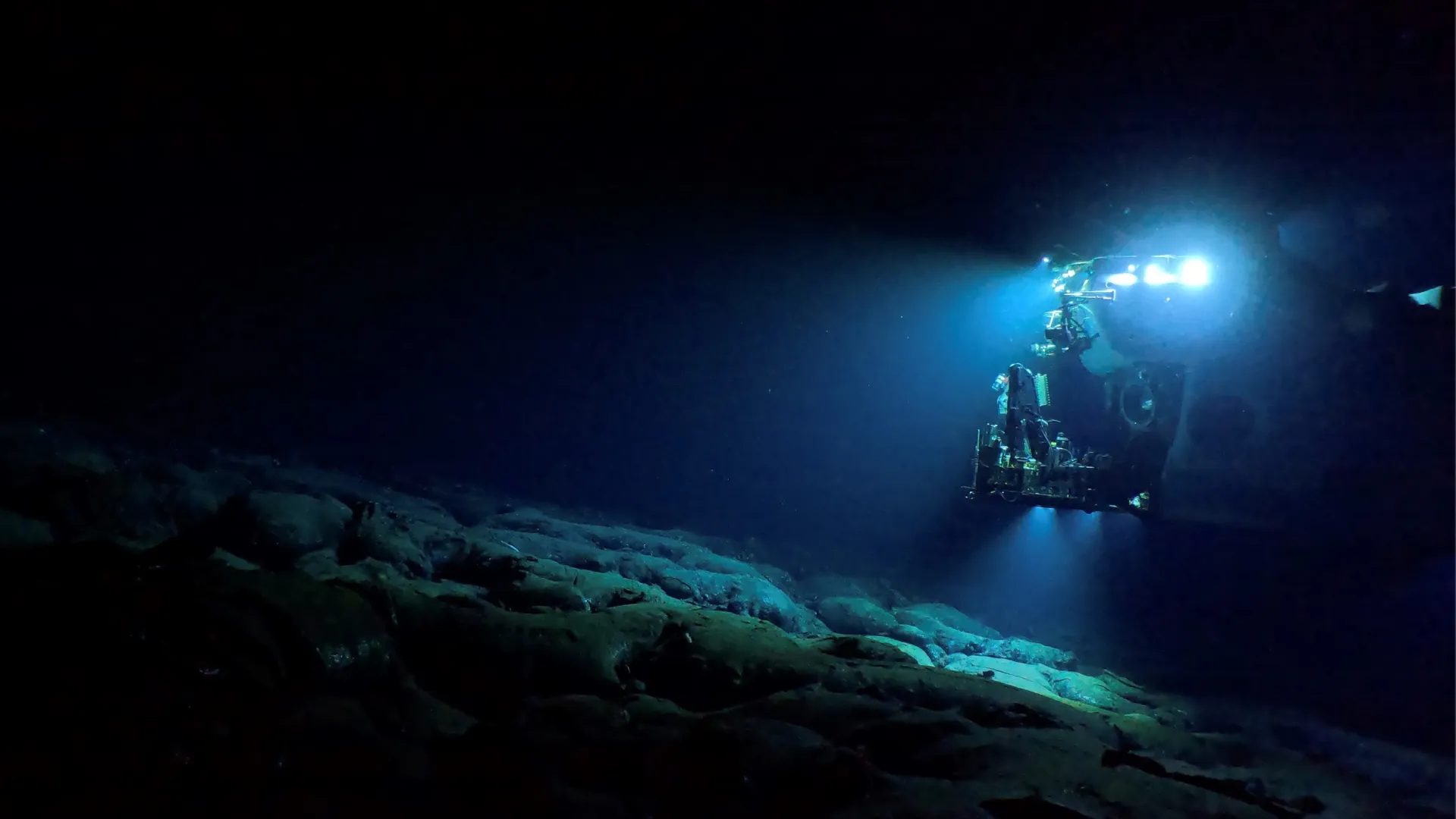 Underwater view of the Alvin submersible lit brightly in dark blue water.