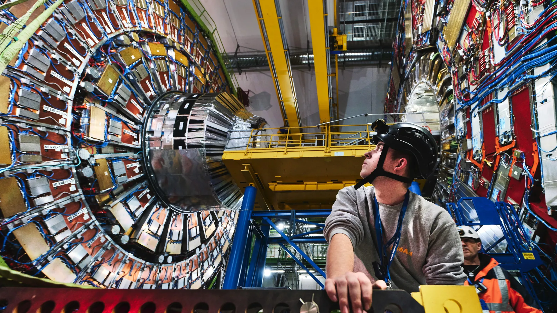 A researcher in a helmet looks up at a large ring of metal devices in the Large Hadron Collider.