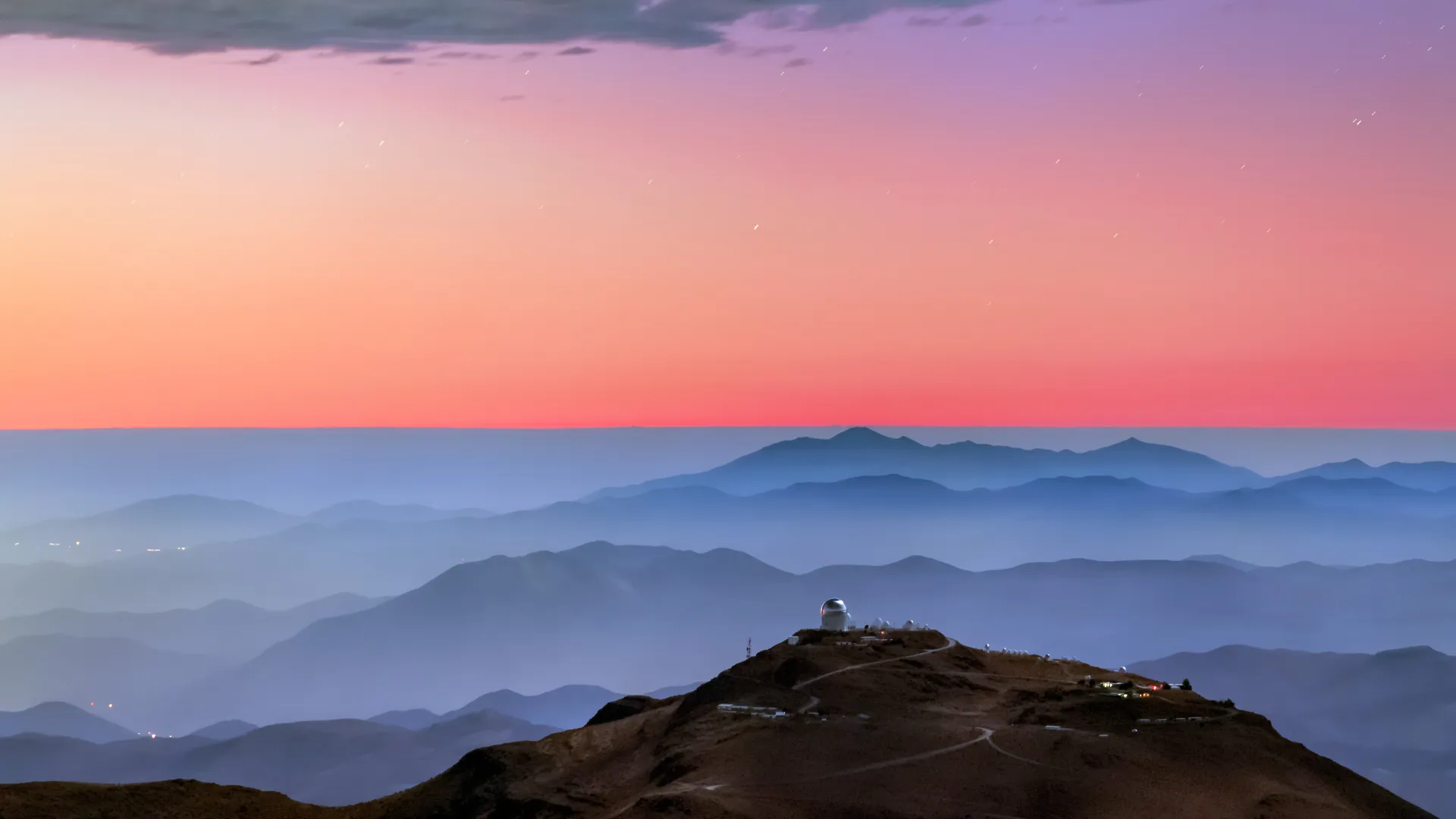 A colorful sunset and layers of blue mountaintops backdrops the Cerro Tololo Inter-American Observatory. A small profile image of the Víctor M. Blanco 4-meter Telescope is visible atop Cerro Tololo itself.