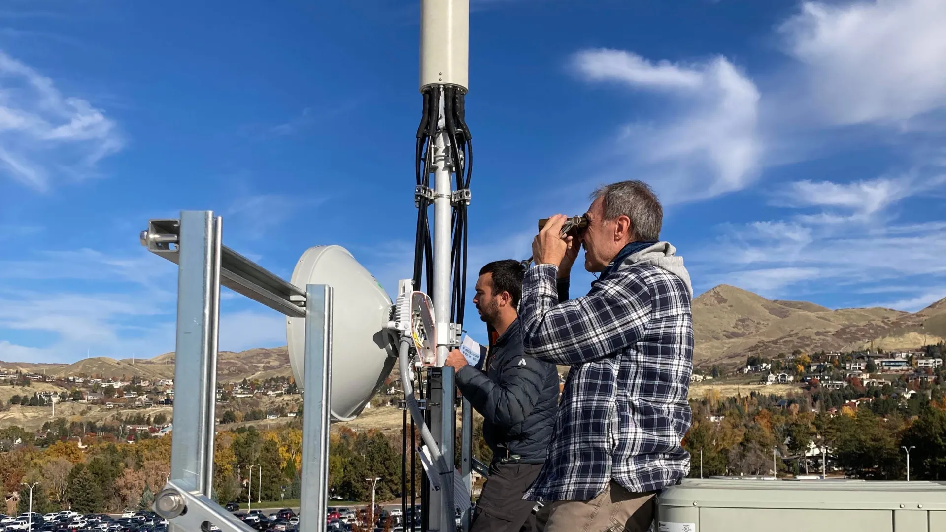 Two people, one with binoculars, stand on some equipment in front of a hillside under a bright blue sky.