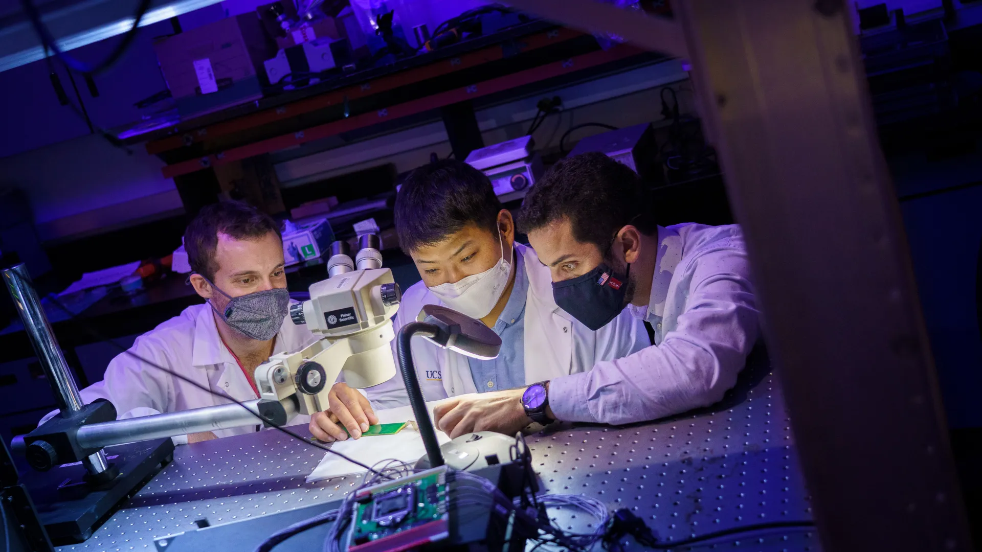 Three researchers in lab coats look at a small sensor.