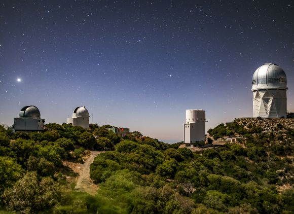 Nighttime scene of a hilly area with five buildings, including observatories, against a background of a star-filled sky.