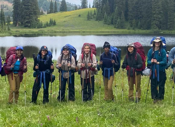 A line of smiling students with large backpacks and hiking poles posing in a meadow in front of a lake.