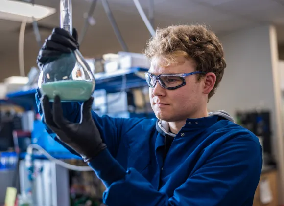 A scientist wearing protective lab gear holds up a flask containing a blue liquid.