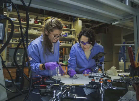 Two women in blue lab coats and purple gloves bend over a lab bench.
