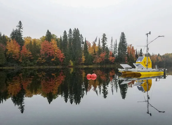 A yellow platform with solar panels and other equipment on it floating on a still lake with trees showing fall colors in the background.