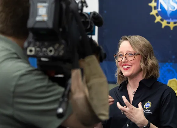 A scientist being filmed by a news camera while speaking in front of an NSF banner.