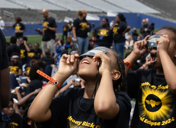 Two kids in matching eclipse t-shirts look up at the sky through paper eclipse glasses.