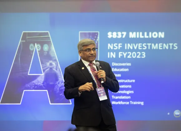 The NSF Director speaks into a microphone against a background display that states "AI: $837 million: NSF investments in FY2023"