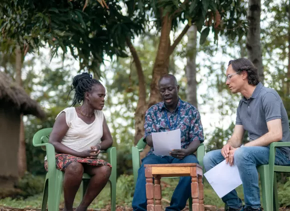 Researchers sitting in plastic chairs surrounded by trees outside a house in Kenya.
