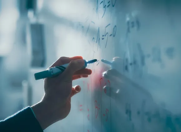 Close-up image of a hand writing equations on a white board.