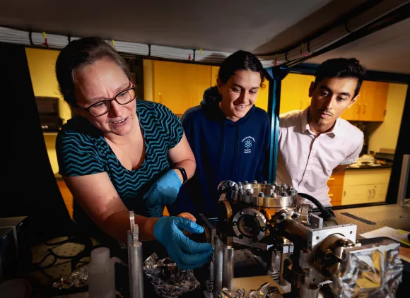 Three people leaning in to inspect a series of device components with various chambers metallic segments.