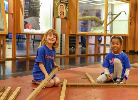 Two kids sitting on the floor in a science center building a wooden structure.