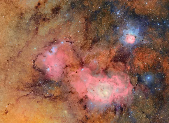 Against a background dense with glowing orange and golden stars, two nebulas appear as pinkish clouds of gas and dust.