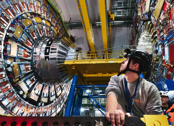 A researcher in a helmet looks up at a large ring of metal devices in the Large Hadron Collider.