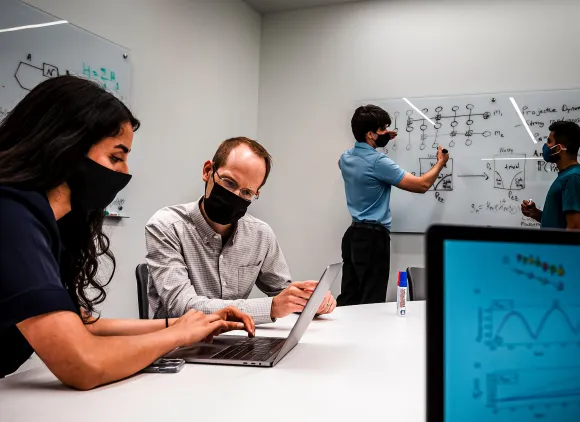 Student with scientist sitting in front of laptop together while two students in the background diagram systems at the white board