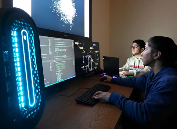 Two students work on computers while looking at a brightly lit screen.