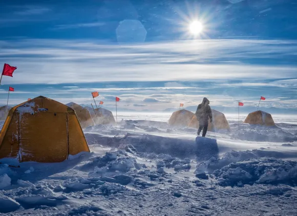 A person walks amidst yellow tents in snowy windy landscape.