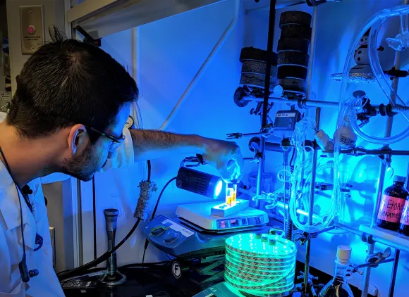 A chemist manipulates equipment in a fume hood lit with blue light.