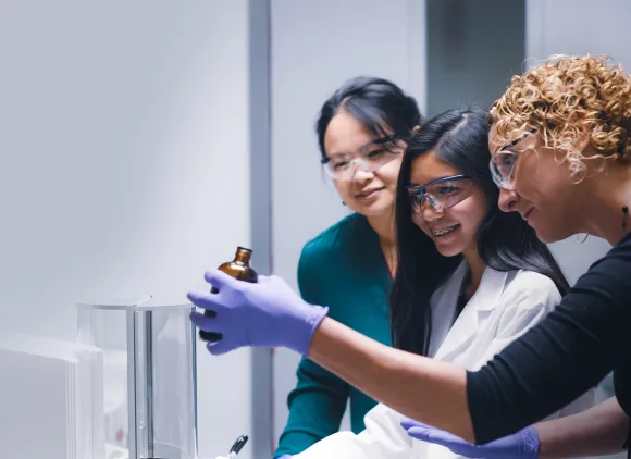 Interns in lab coats and protective eyewear looking at bottled sample held by scientist.