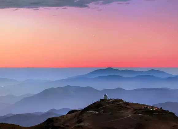 A colorful sunset and layers of blue mountaintops backdrops the Cerro Tololo Inter-American Observatory. A small profile image of the Víctor M. Blanco 4-meter Telescope is visible atop Cerro Tololo itself.