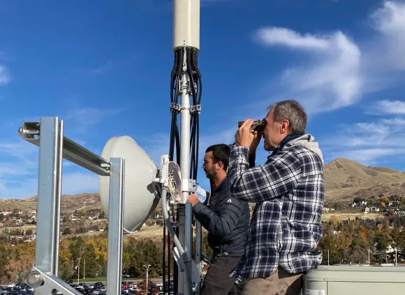 Two people, one with binoculars, stand on some equipment in front of a hillside under a bright blue sky.